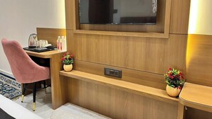 A hotel room with a wooden desk, a pink chair, a television mounted on the wall, and two small potted plants. - Hotel prime bliss (Kanpur)