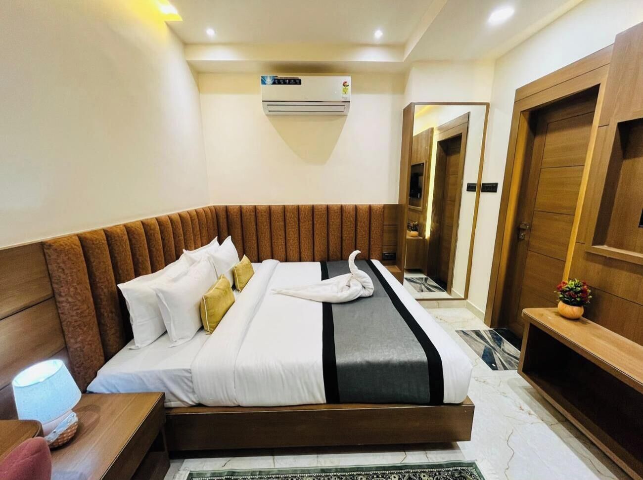 A hotel room with a large bed, a bedside table with a lamp, a wooden headboard, a mirror, and a small plant.