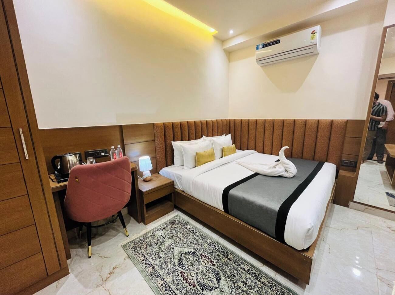 Une chambre d’hôtel avec une tête de lit en bois, un lit avec une literie blanche, un bureau avec une chaise, une petite table avec une lampe et un tapis à motifs.