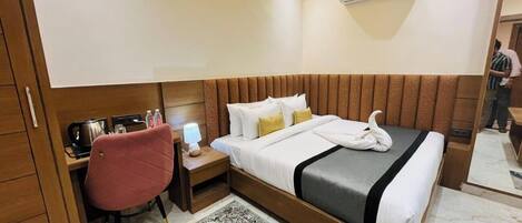 A hotel room with a wooden headboard, a bed with white bedding, a desk with a chair, a small table with a lamp, and a patterned rug.