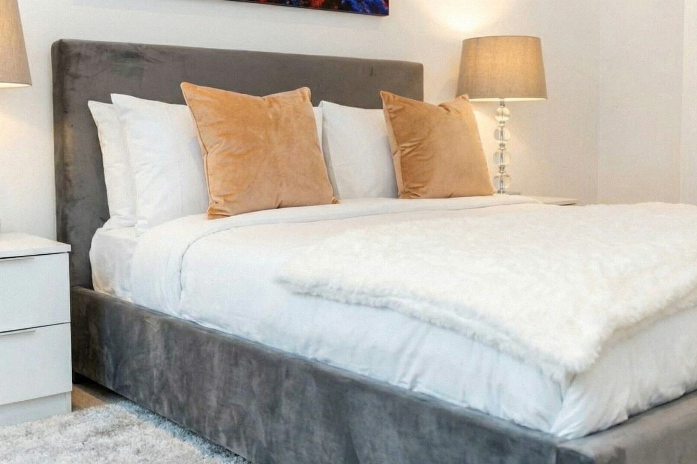 A neatly made bed with a grey headboard and white bedding, a bedside lamp, and a white nightstand.