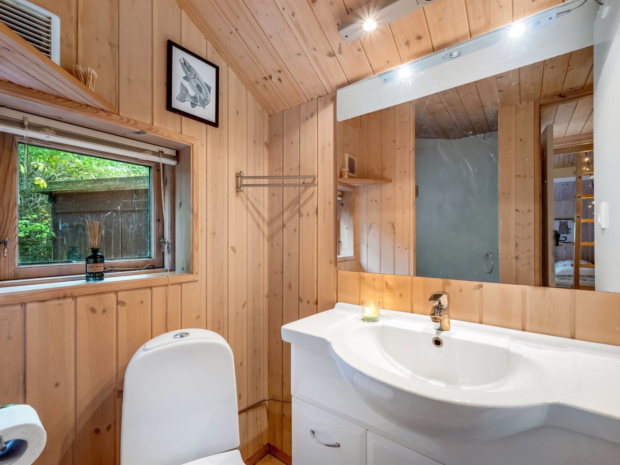 A wooden-paneled bathroom with a large mirror, a white sink, and a window with a view of greenery.