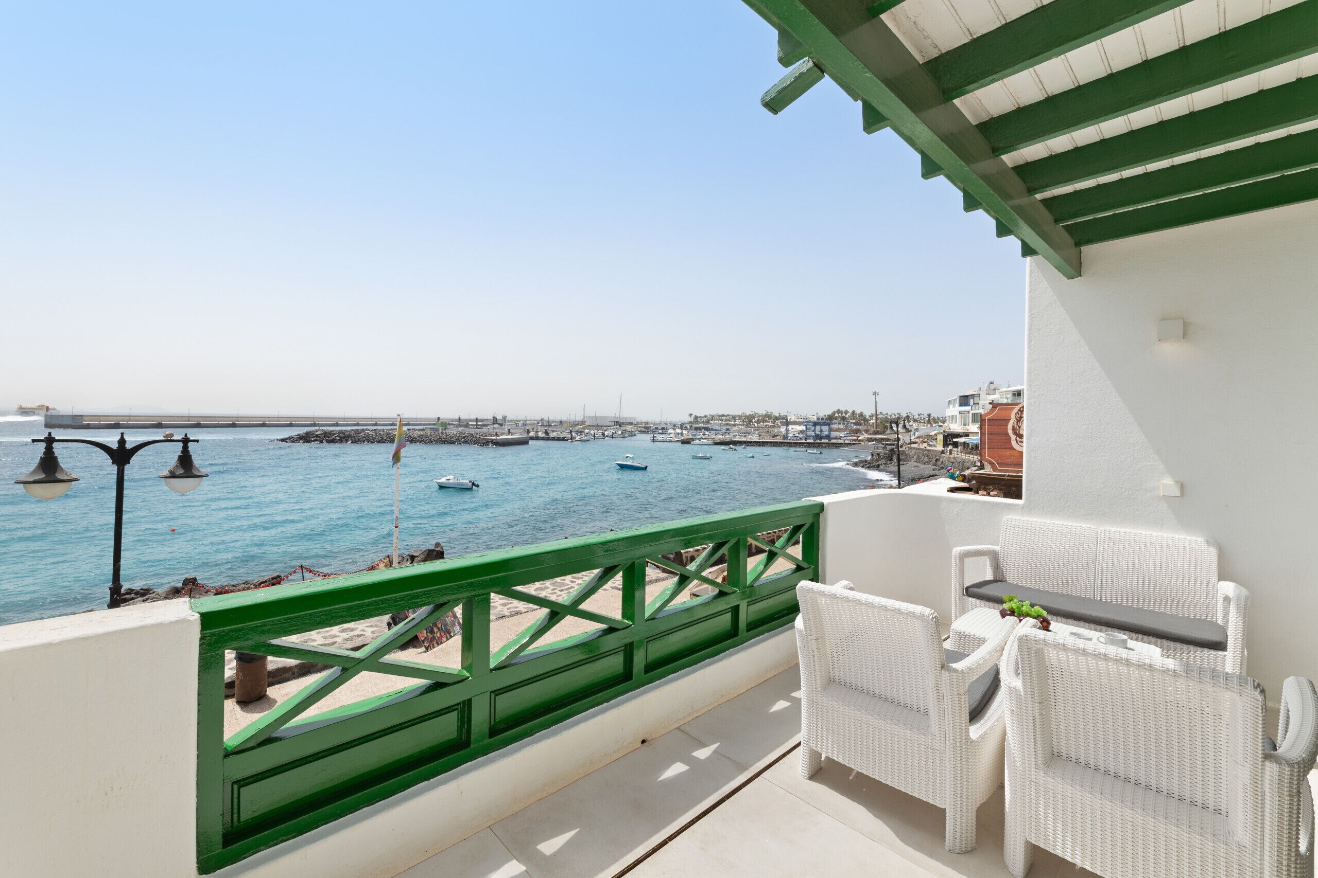 A balcony with white wicker furniture, a green railing, and a view of the sea with boats.
