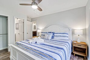 A bedroom with a bed, a ceiling fan, a nightstand, and a door leading to another room. - Beach Bliss Villa with Scenic Ocean Views (Hilton Head Island)