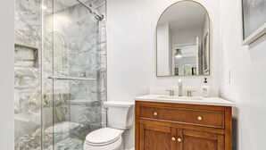 Een badkamer met een douchecabine van glas, een wit toilet, een houten wastafelmeubel met spiegel en een witte wasbak.