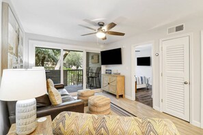 A modern living room with a sofa, ottomans, a lamp, a ceiling fan, and a TV mounted on the wall.