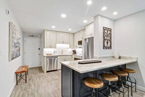 A modern kitchen with white cabinets, a stainless steel dishwasher, a large island with stools, and a refrigerator.
