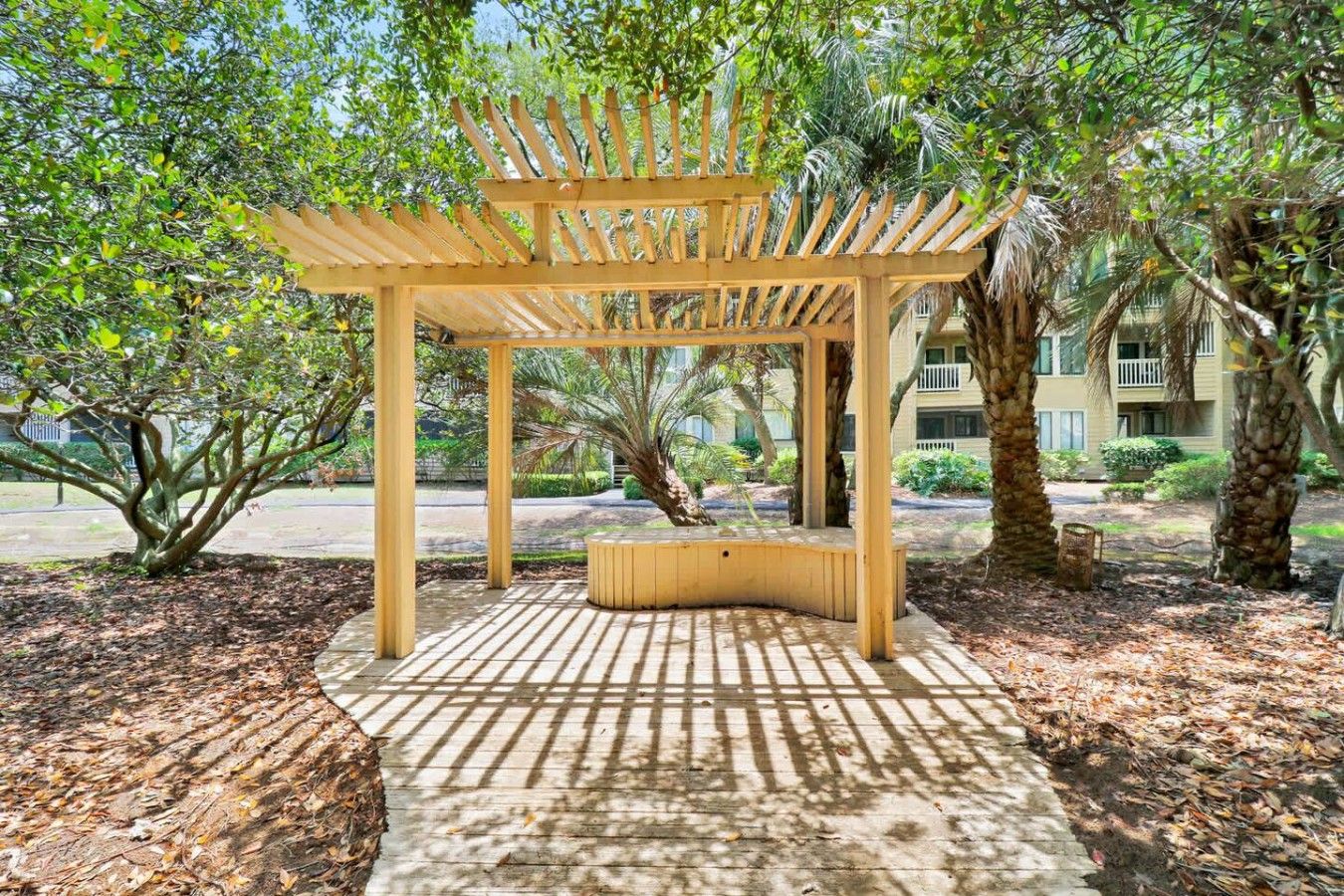 A wooden pergola structure with a bench in a landscaped area with trees and a building in the background.