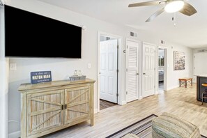 A hallway with a mounted television, a wooden cabinet with a sign, white doors, and a ceiling fan. - Beach Bliss Villa with Scenic Ocean Views (Hilton Head Island)