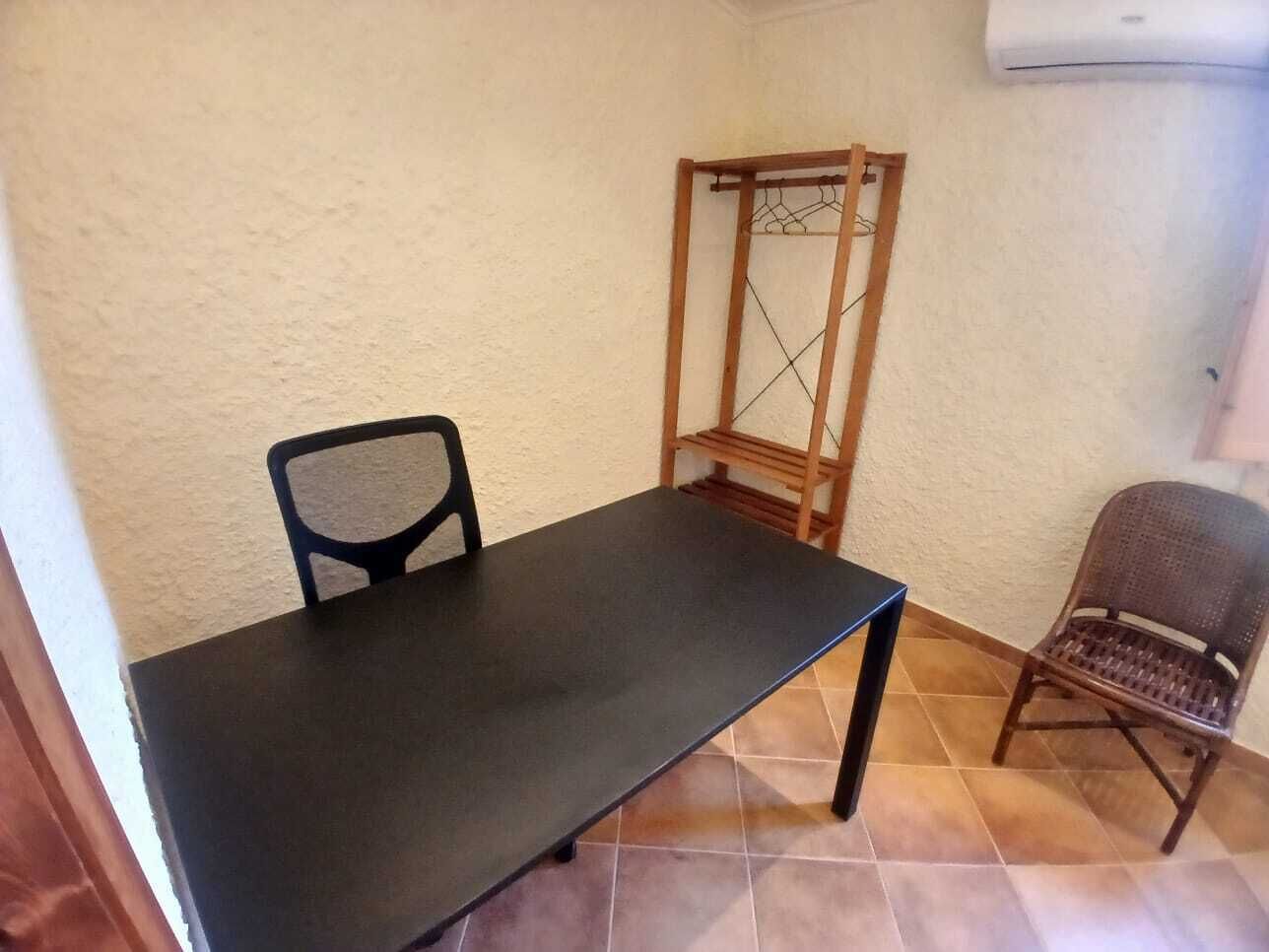 Una habitación con una mesa oscura, dos sillas, un perchero de madera y suelo de baldosas.