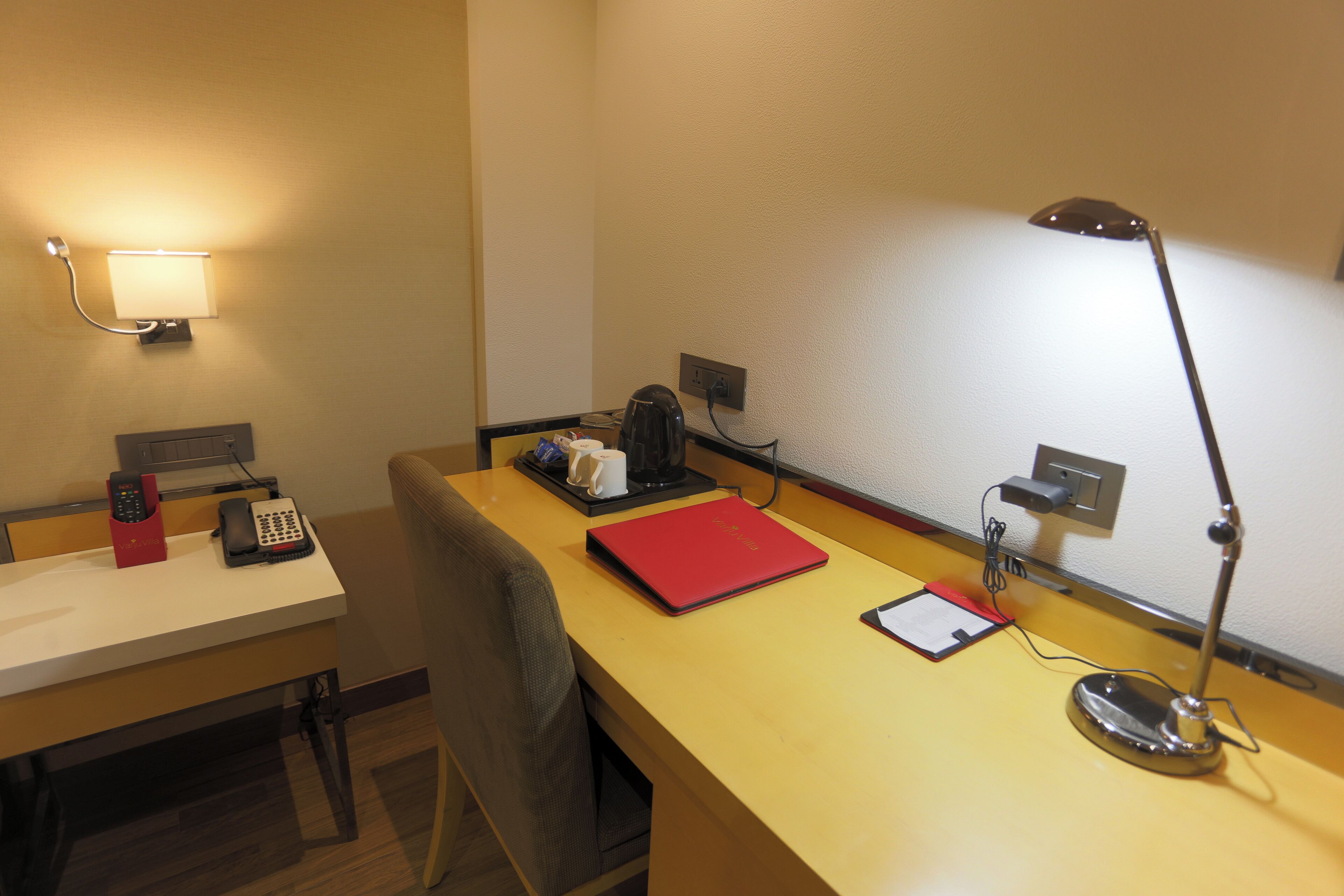 A hotel room with a desk, a lamp, a phone, a red folder, a coffee maker, and a wall socket.