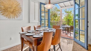 Uma sala de jantar com mesa posta para quatro pessoas, um ventilador de teto e uma janela grande com vista para um jardim.