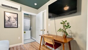 Une pièce avec une table à manger en bois, une chaise rembourrée de couleur rouge, un téléviseur fixé au mur, une plante en pot et un tableau encadré accroché au mur.