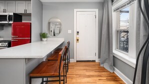 Une cuisine moderne avec des meubles blancs, un réfrigérateur rouge, un banc en bois et une grande fenêtre avec des rideaux.