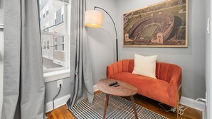 Une pièce avec un canapé gris, une petite table ronde, un lampadaire et un grand tableau encadré au mur.