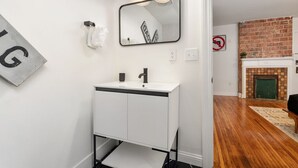A modern bathroom with a white vanity, a large mirror, and a brick fireplace in the background.