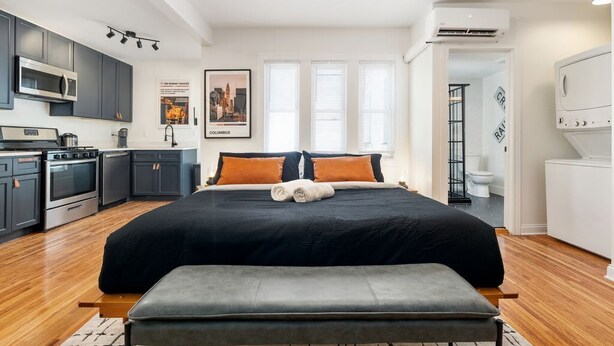 A modern bedroom with a large bed, a bench, a kitchenette, and a bathroom visible through an open door.