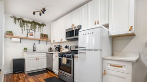 A modern kitchen with white cabinetry, a stainless steel refrigerator, a microwave, and a stove. There are wooden shelves above the counter and a small black trash bin to the left. - Modern Loft In the Heart of OSU Campus (Columbus)