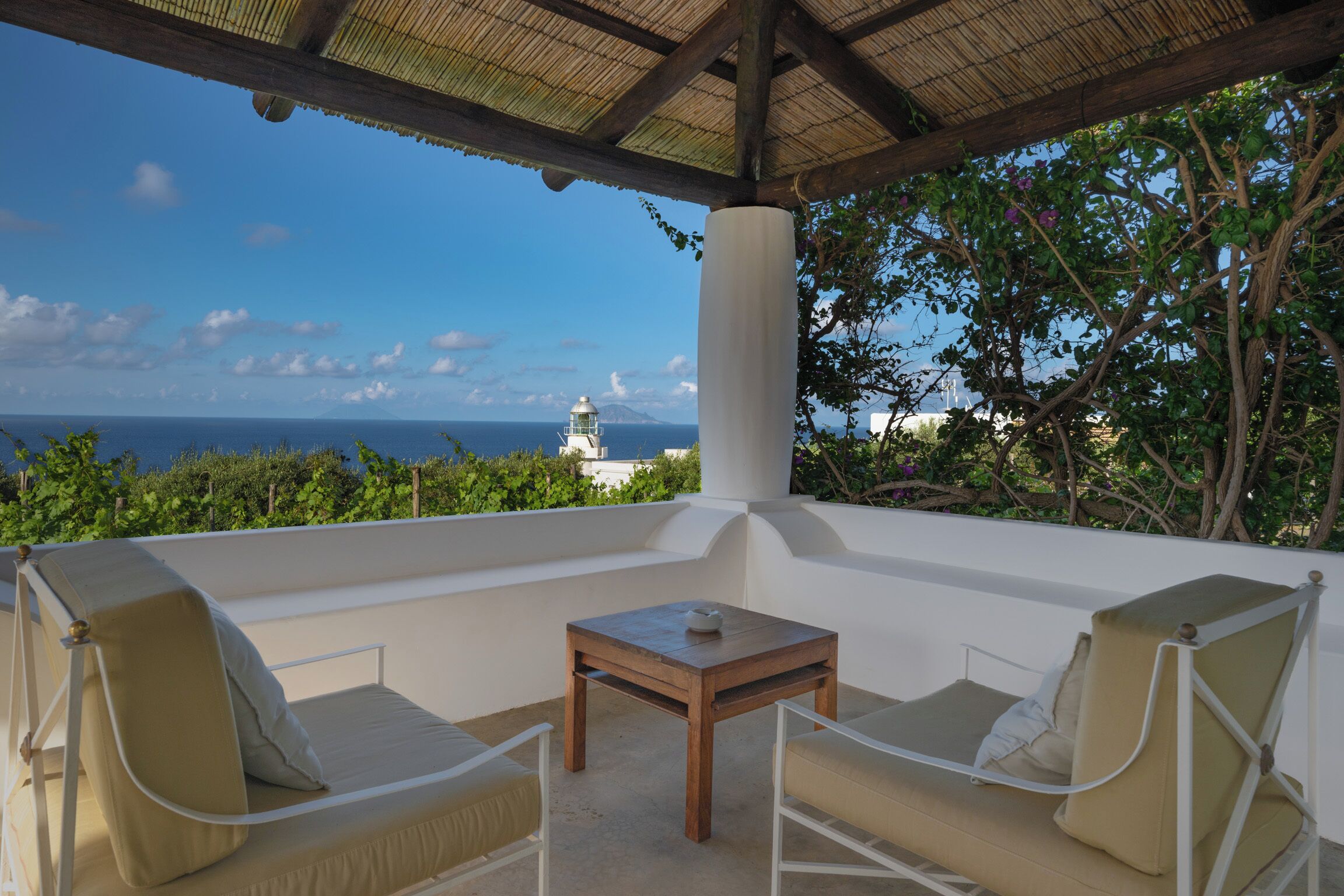 A covered outdoor seating area with a table and chairs, overlooking a scenic view of the ocean and a lighthouse.