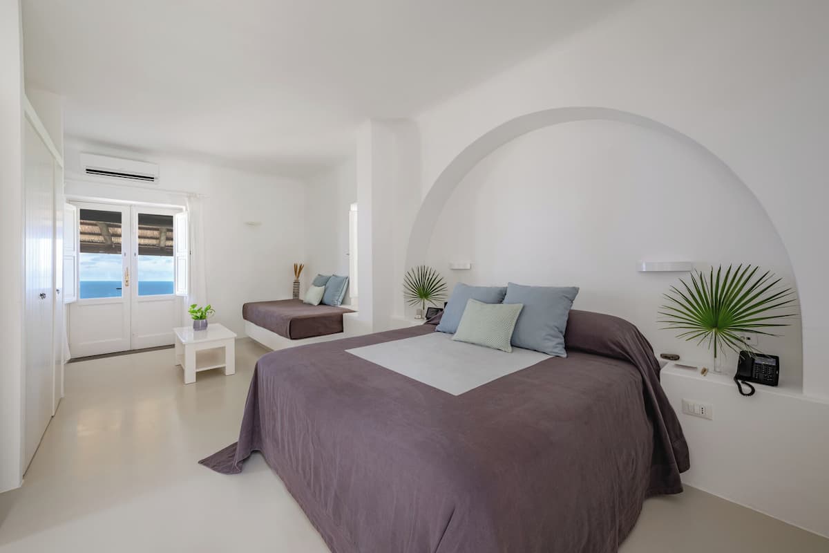 Ein Schlafzimmer mit einem großen Bett, einer kleinen Sitzecke, einem Fenster mit Ausblick und einer Pflanze auf dem Nachttisch.