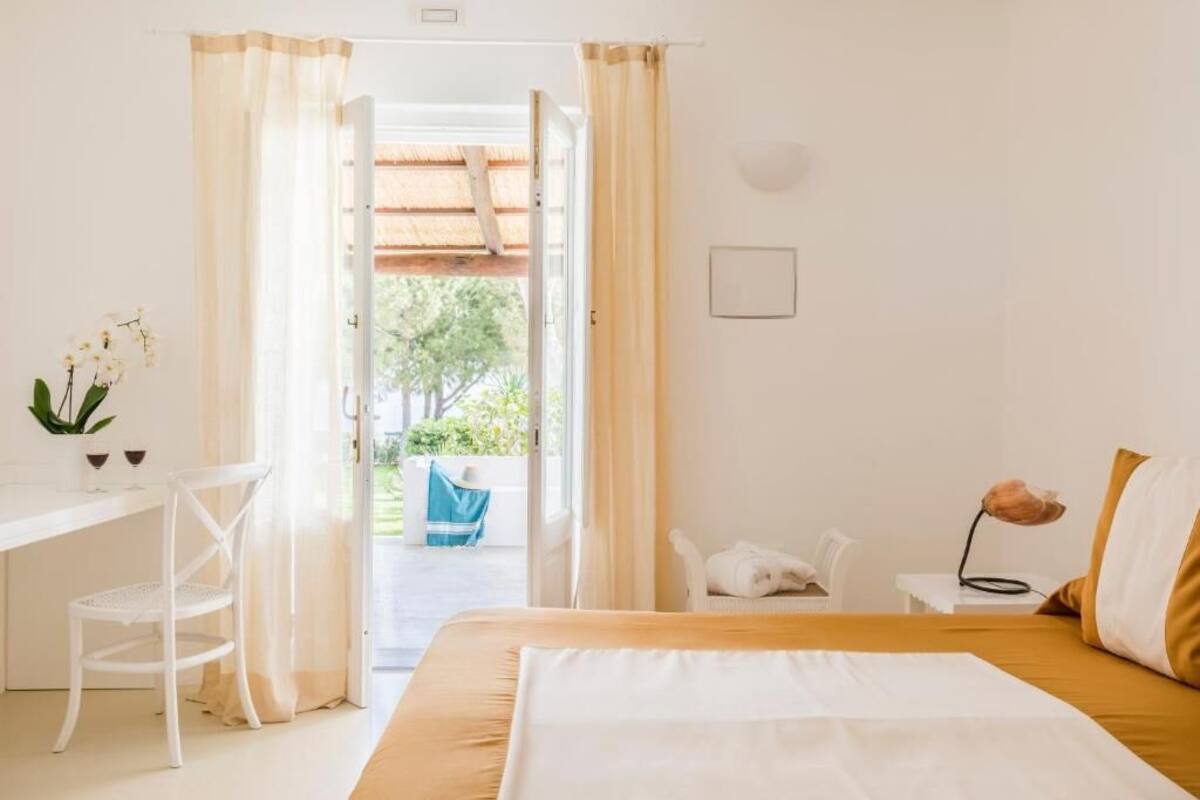 Ein Schlafzimmer mit Bett, Schreibtisch und Stuhl. Ein Fenster mit Vorhängen bietet Blick ins Freie, und ein kleiner Tisch ist mit einer Lampe bestückt.