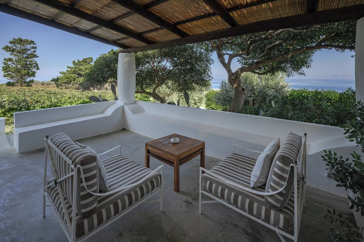Ein Sitzbereich im Freien mit zwei gestreiften Sofas und einem kleinen Tisch, mit Blick auf eine malerische Landschaft mit Bäumen und dem Meer.