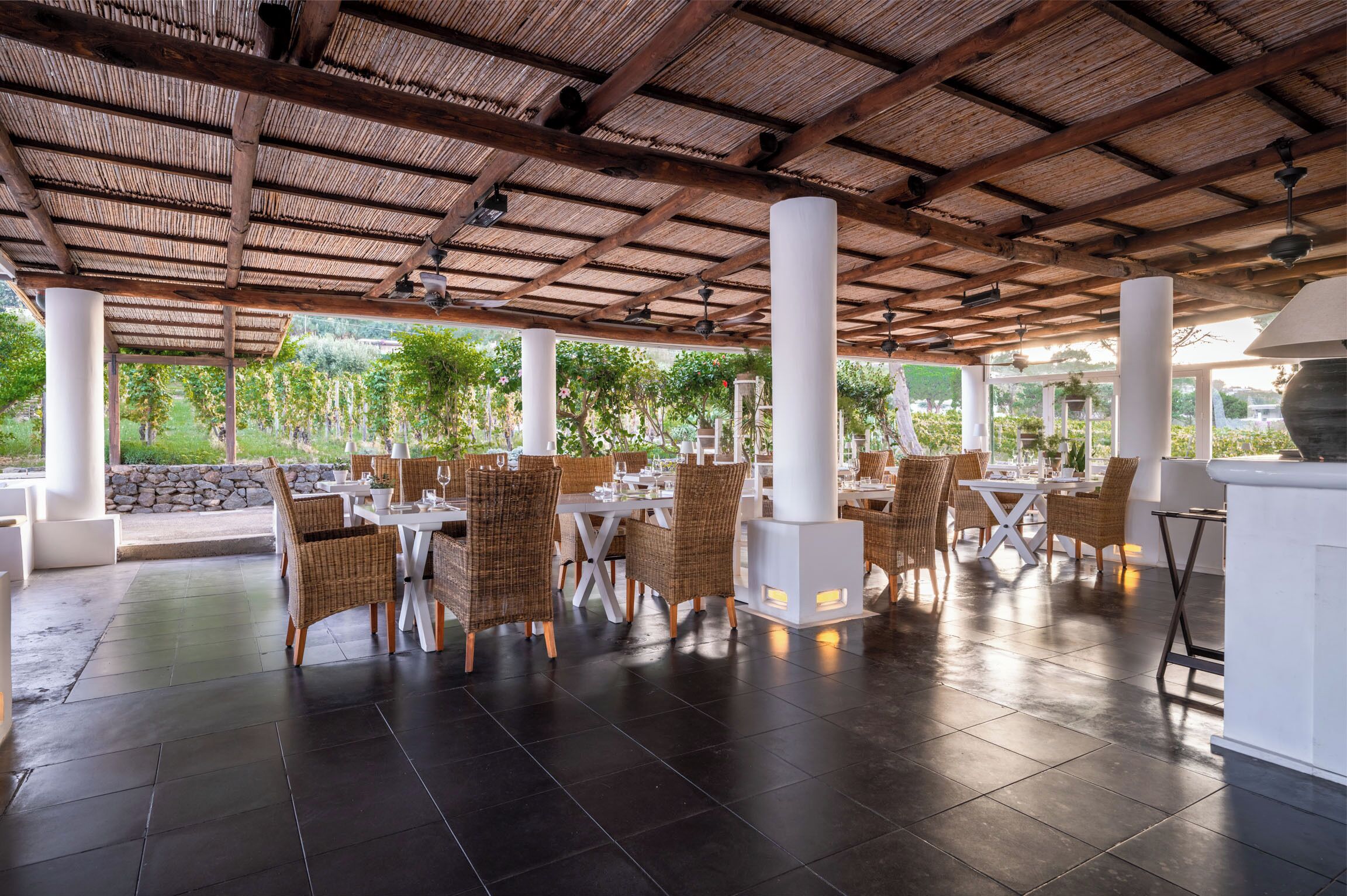 An outdoor dining area with wicker chairs and tables, a thatched roof, and a view of greenery.