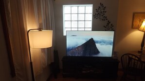 Un salon avec un téléviseur à écran plat diffusant un paysage de montagne, un lampadaire, un fauteuil et une fenêtre avec des stores.
