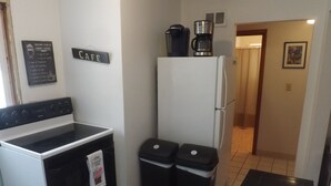 Une petite cuisine équipée d’une cuisinière, d’un réfrigérateur et de deux poubelles. Une machine à café est posée sur le réfrigérateur, et une pancarte sur le mur indique « CAFÉ ».