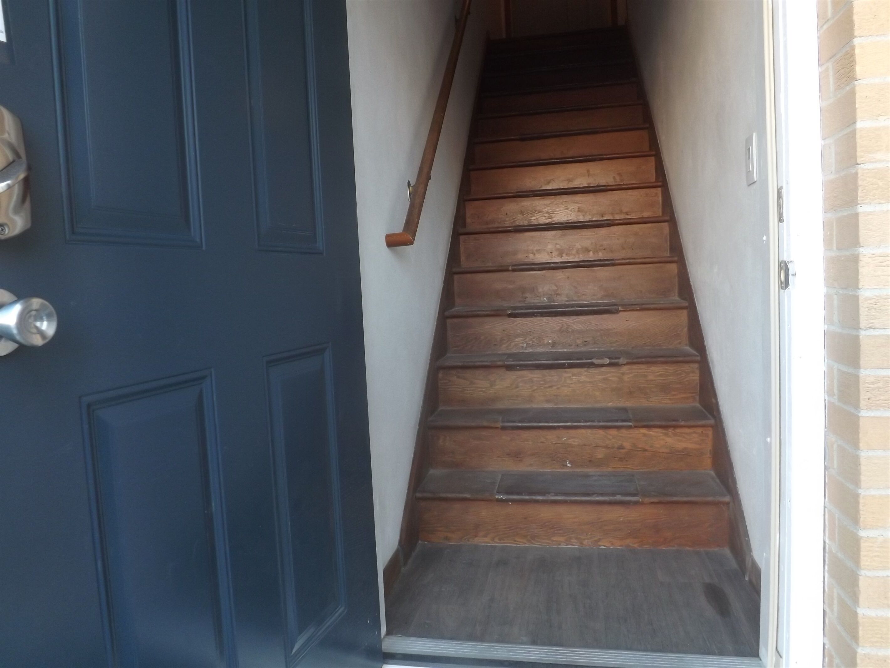 A wooden staircase with a dark blue door on the left and a brick wall on the right.