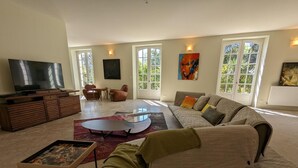 A spacious living room with a large sofa, a coffee table, and a TV on a wooden cabinet. There are large windows with a view of greenery outside. - Large house with private pool, wifi, pets allowed for 15 guests. (Pranzac)