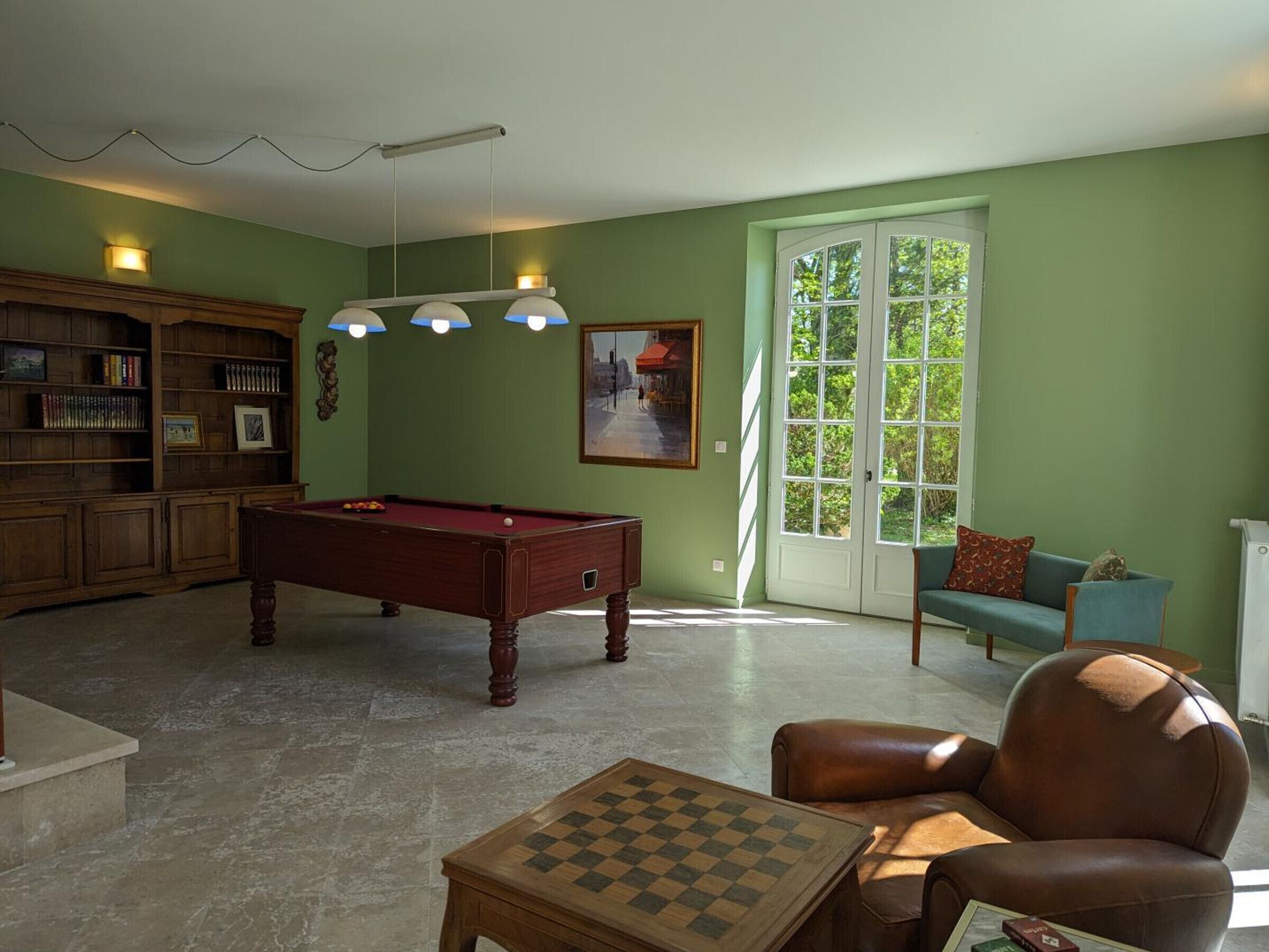 Una sala de estar con mesa de billar, estantería, sofá y ventana con vista a la vegetación.