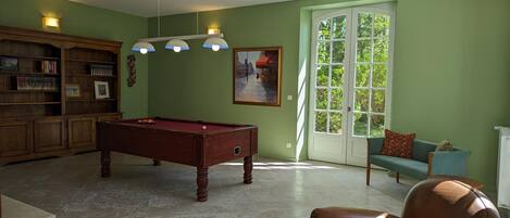 Una sala de estar con mesa de billar, estantería, sofá y ventana con vista a la vegetación.