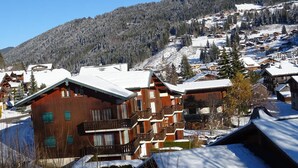 A wooden chalet-style building with multiple balconies, surrounded by snow-covered houses and a mountainous backdrop. - 3-room Apartment 5 guests - South Terrace, Garage, WiFi, Near shops (Les Gets)
