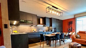 A modern kitchen with a central island, dining area with chairs, and a seating area with a sofa and a coffee table. - 3-room Apartment 5 guests - South Terrace, Garage, WiFi, Near shops (Les Gets)