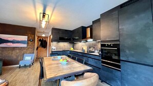 A modern kitchen with dark cabinets, a wooden dining table, and a large window. - 3-room Apartment 5 guests - South Terrace, Garage, WiFi, Near shops (Les Gets)