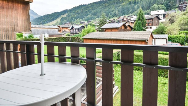 A wooden deck with a round table, overlooking a village with traditional houses and a forested mountain backdrop.