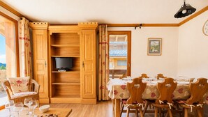 A dining area with a wooden table and chairs, a television on a shelf, and a view of a building through an open door. - Village center apartment with pool and terrace (Notre-Dame-de-Bellecombe)