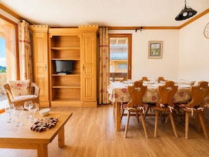 Un comedor con una mesa y sillas de madera, un televisor en un estante y vistas de un edificio a través de una puerta abierta.