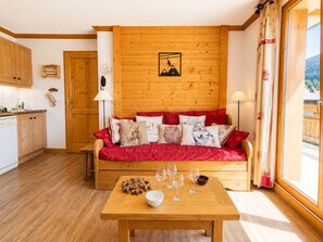 A cozy room with a wooden sofa, a dining table with glasses and a bowl, and a kitchen area with wooden cabinets.