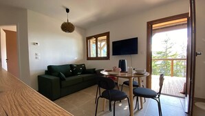 A modern living room with a sectional sofa, a round dining table with chairs, a flat-screen TV mounted on the wall, and a balcony with a wooden deck. - Modern 2-room apartment + cabin, 4 persons, parking (Cohennoz)