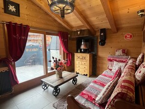 A cozy wooden cabin with a sofa, a wooden table with a flower vase, a fireplace, and a view of the outside.