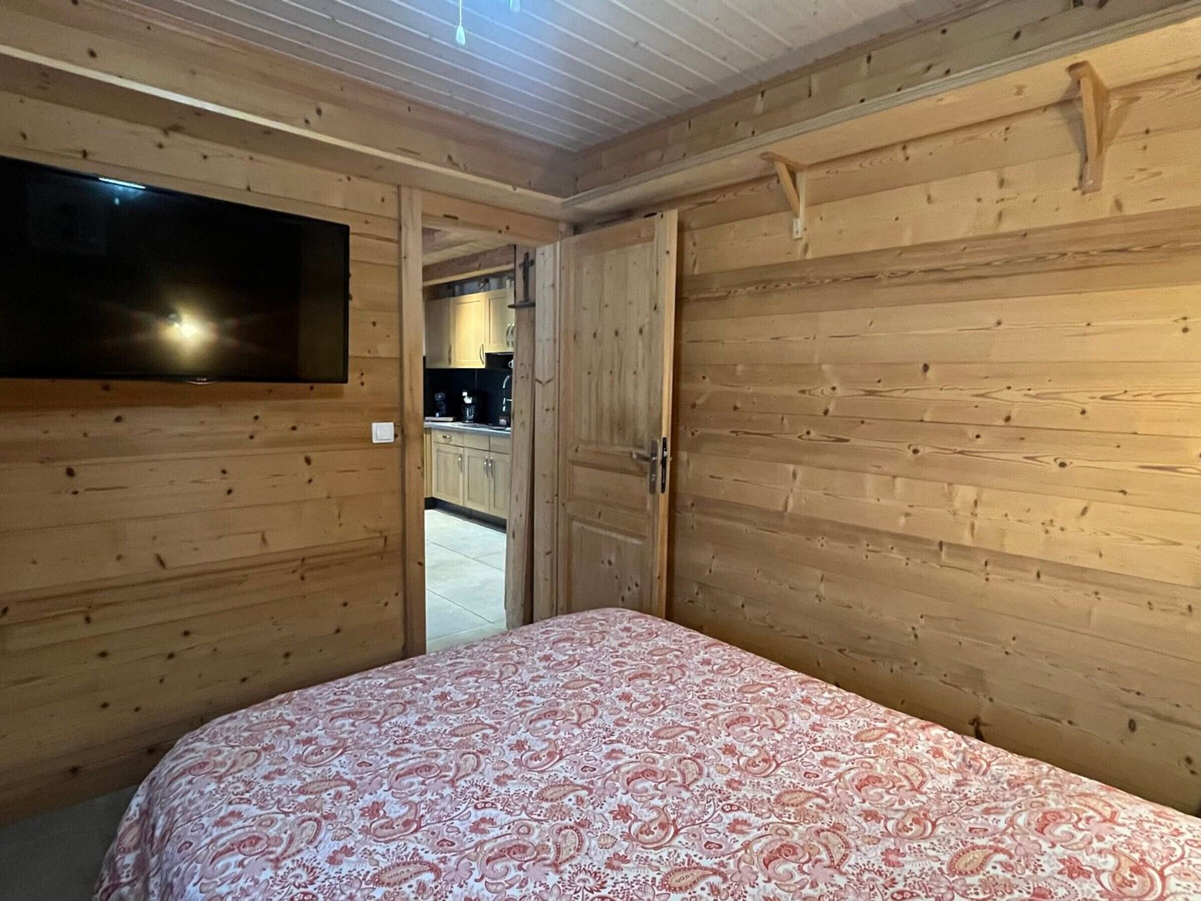 Une pièce aux murs lambrissés, comprenant un lit, un téléviseur fixé au mur et une porte donnant sur un coin cuisine.