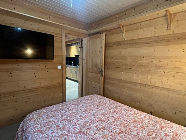 A wooden-paneled room with a bed, a TV mounted on the wall, and a door leading to a kitchen area.