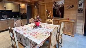 A cozy wooden interior with a dining table set for four, surrounded by wooden chairs. A kitchen with modern appliances and wooden cabinets is visible in the background. - Châtel: 3-Room Apt, Terrace, 5 Pers, Private Parking (Châtel)