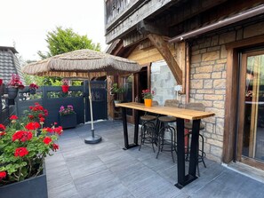 Un espace patio avec une table et des chaises en bois, entouré de plantes en pot et d’un parasol en chaume.