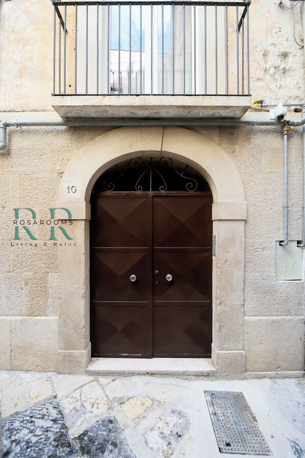 Unas puertas dobles con un arco decorativo encima, un balcón con barandillas de metal y una señal con el texto 'ROSARUM' en la fachada del edificio.