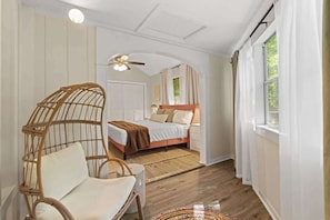 A bedroom with a bed, a wicker chair, a ceiling fan, and a window with curtains.