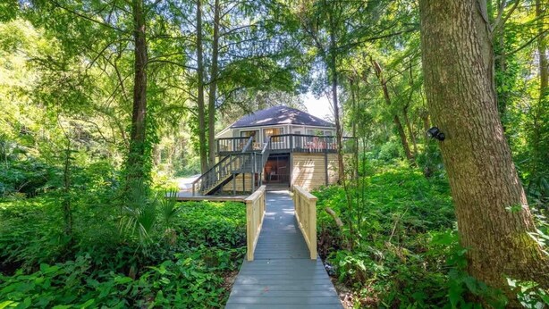 Eine Holzbrücke führt zu einem Haus, umgeben von dichtem Wald.