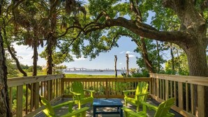 Eine Holzterrasse mit grünen Stühlen und einem Tisch, mit Blick auf ein Gewässer und eine Brücke.